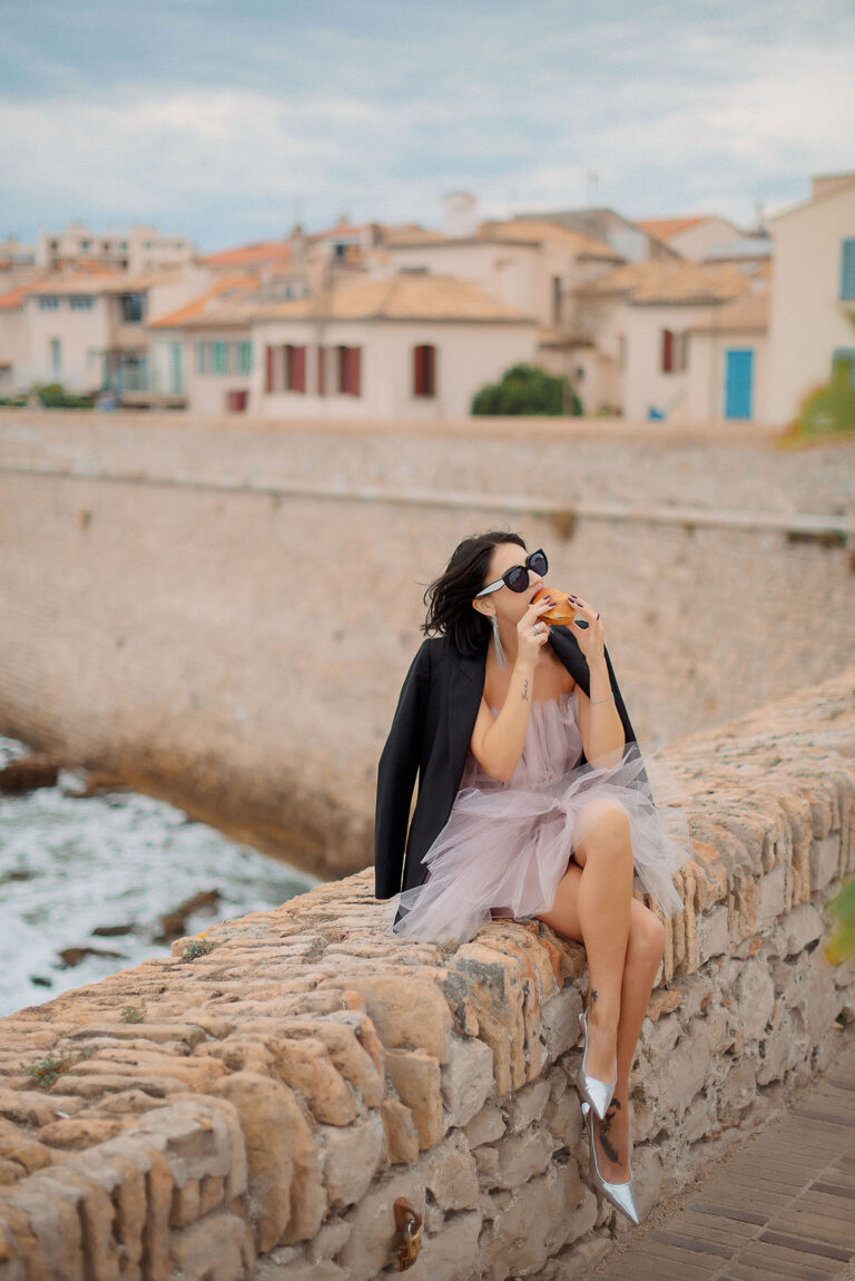 Photographe à Antibes : portrait assis sur un rempart en robe, dégustant un bagel avec vue sur la vieille ville