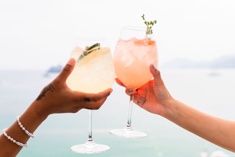 Un toast au coucher du soleil : deux cocktails s’entrechoquent sur fond de mer Méditerranée.