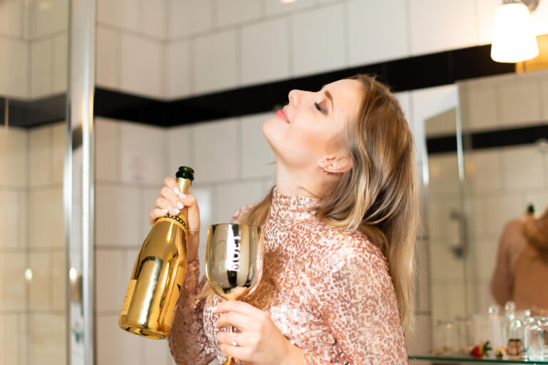 Photographe lifestyle à Nice : portrait d’une fille pétillante avec une coupe de champagne dans une salle de bain chic.