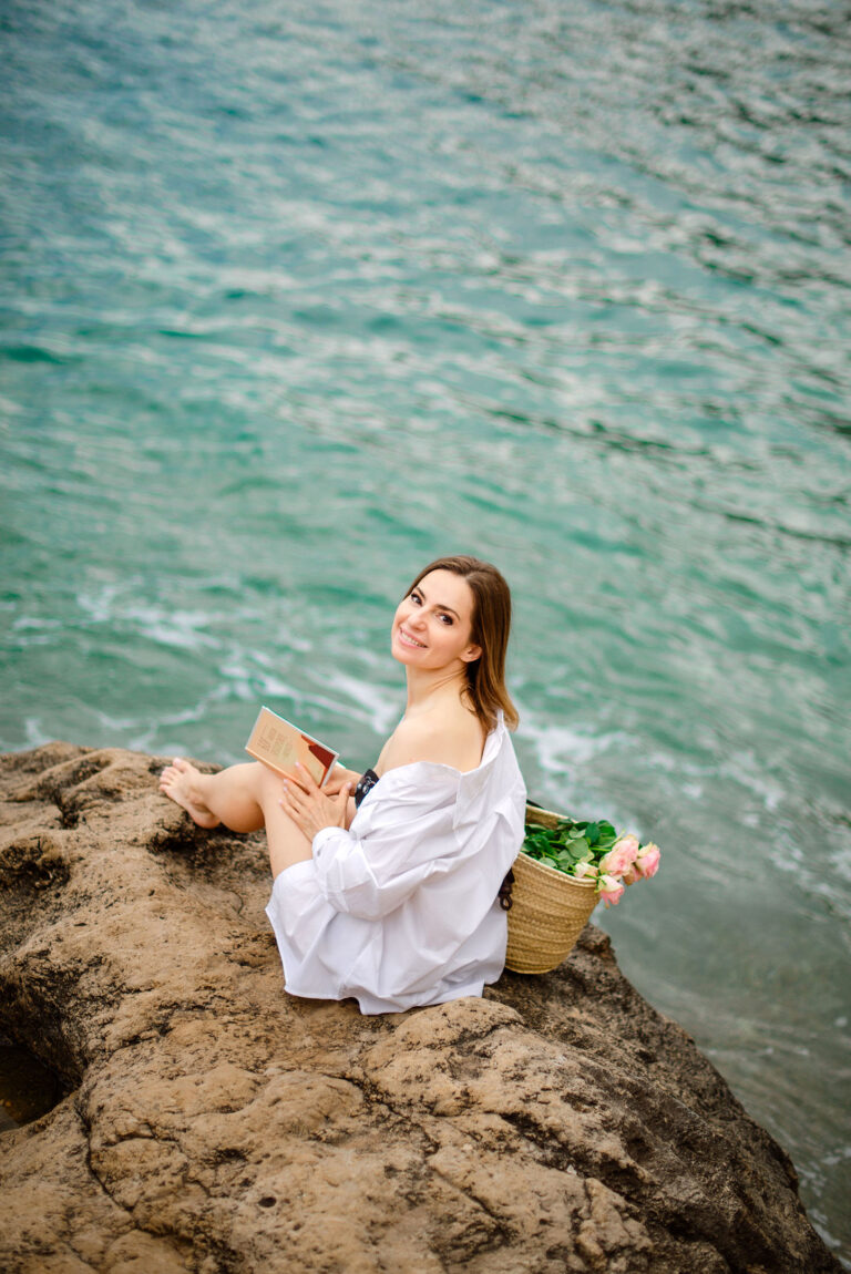 Portrait de femme en maillot et chemise blanche, lisant sur un rocher en bord de mer.