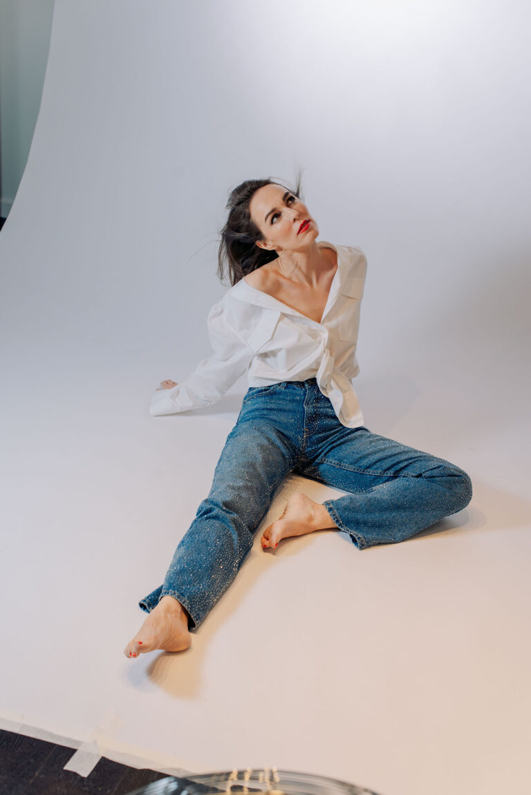 Femme assise dans un décor minimaliste, chemise blanche et regard confiant.