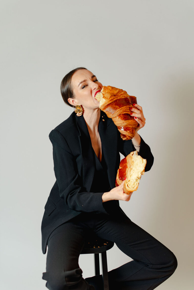 Portrait décalé d’une femme riant en déchirant un croissant géant.