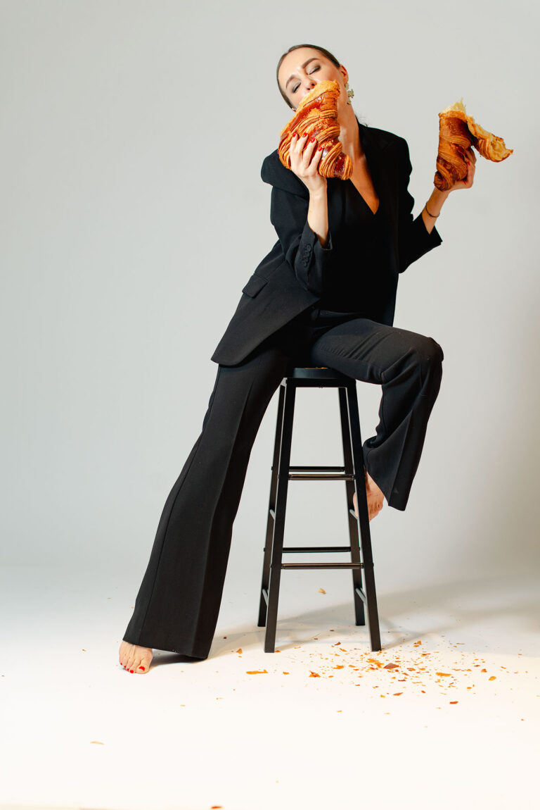 Portrait en costume sur chaise, tenant un croissant géant brisé en deux, ambiance décalée.